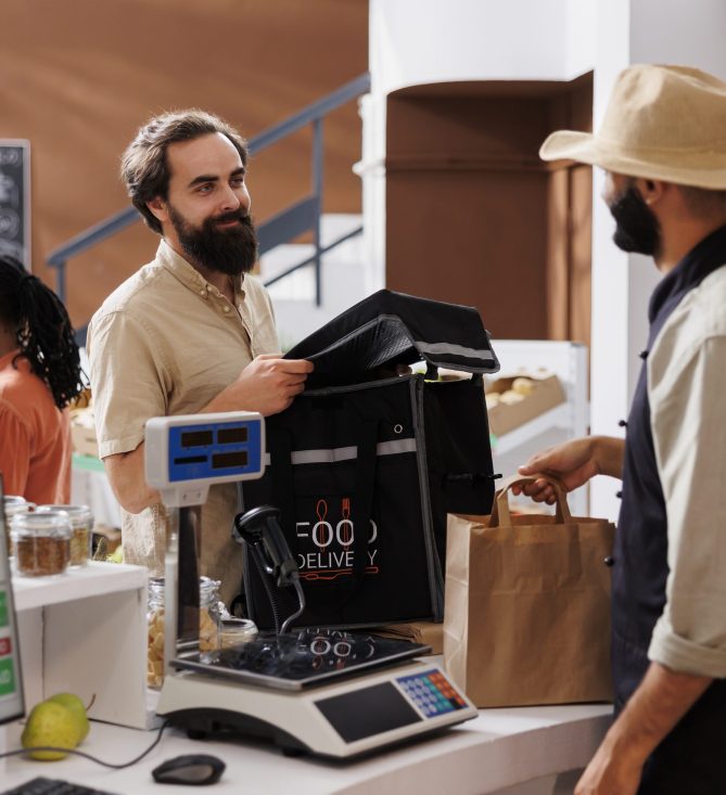 Caucasian male courier arrives at checkout counter ready to receive packaged grocery order from cashier. Food delivery man with backpack retrieves takeaway from vendor at local market.